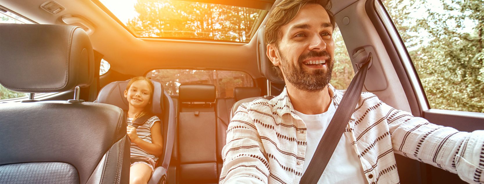 A smiling dad talks to his daughter as they drive the family car through a wooded area.