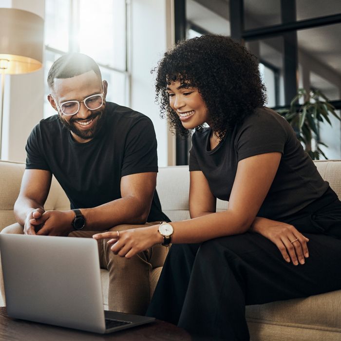 Two Patelco members sitting on a couch and researching options for local resources, both pointing at a laptop and smiling